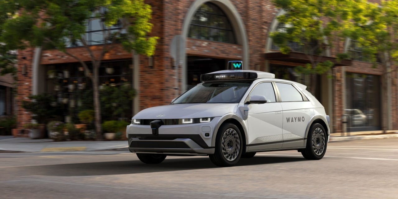 A white Waymo-branded Hyundai Ioniq 5 on a brick-front city street.