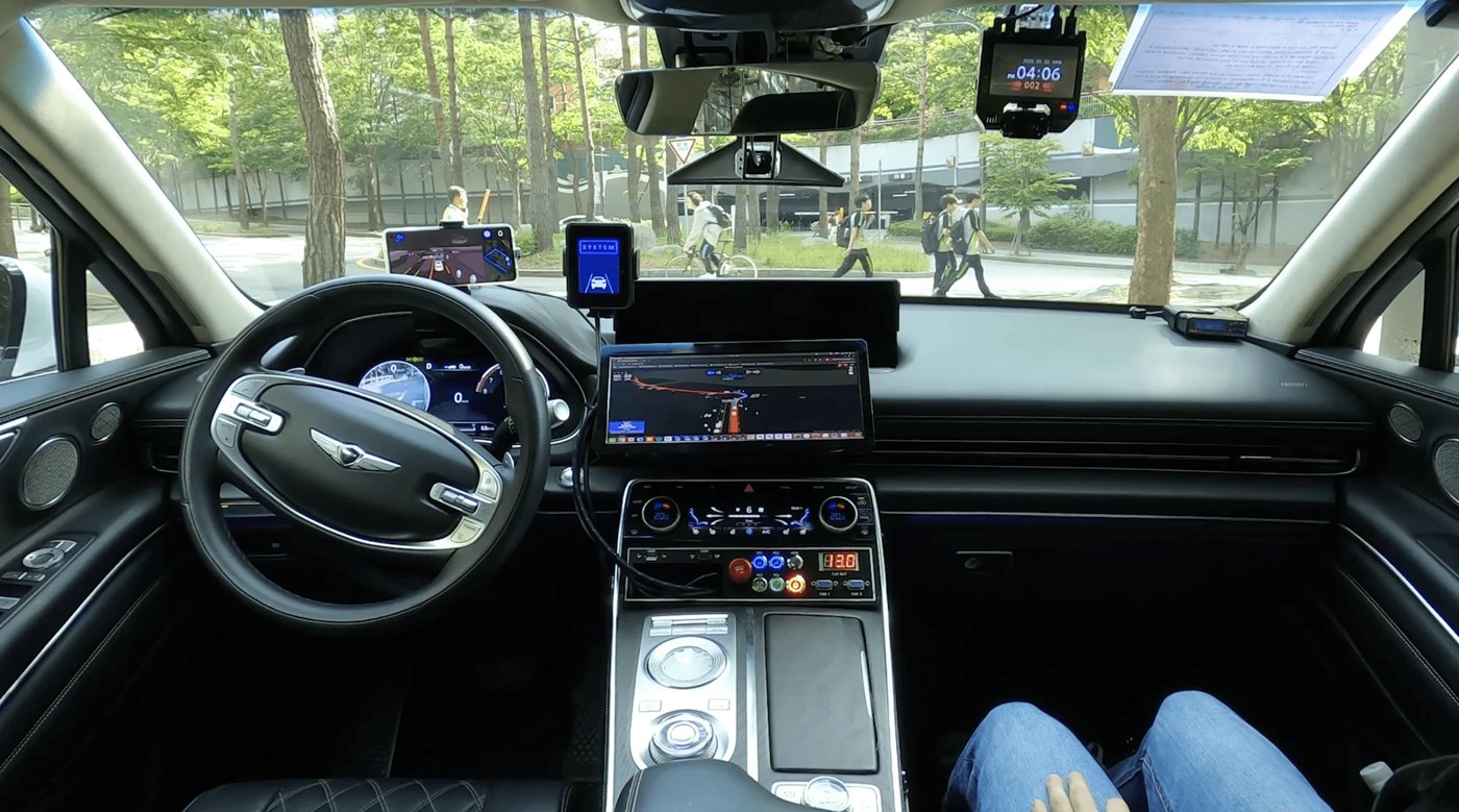 The driver's seat of a RideFlux autonomous Genesis SUV sits empty as the car drives itself past pedestrians, with monitoring screens mounted on the dashboard. Company photo.