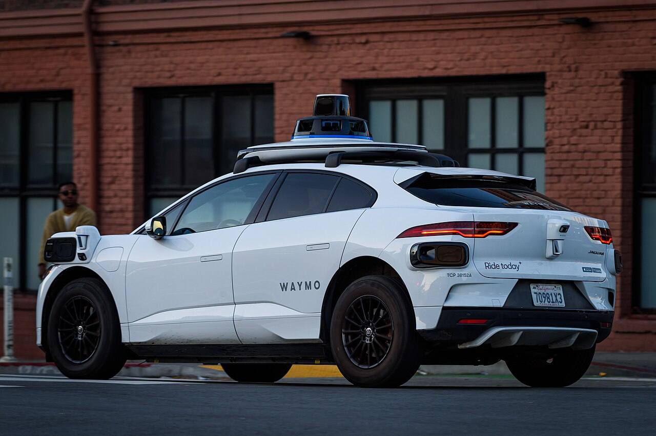 Rear quarter view of a Waymo Jaguar I-PACE, illustrative U.S. street scene.