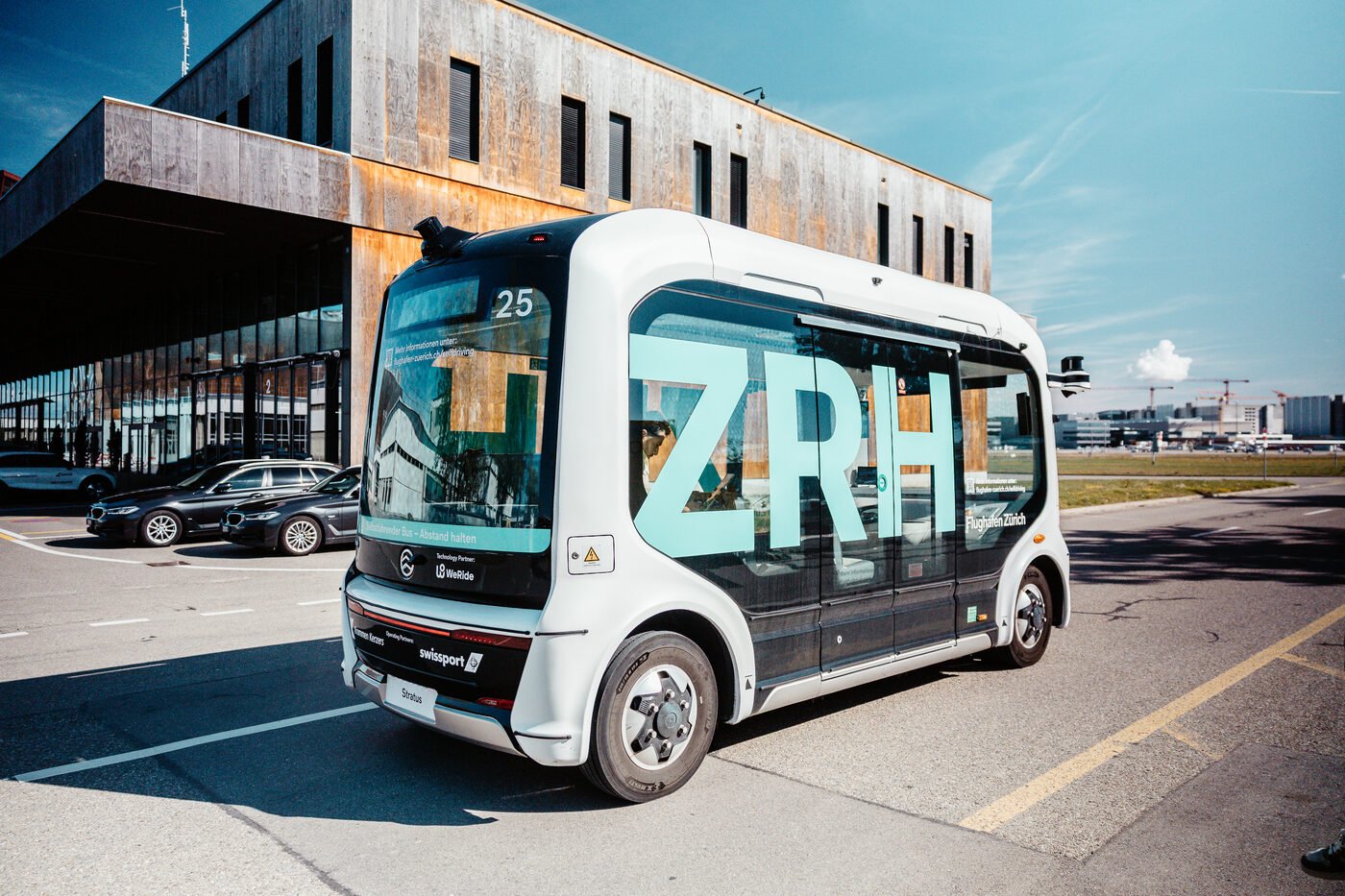 A white WeRide Robobus on the Zurich Airport airfield, with the airport terminal and Swiss aircraft in the background.