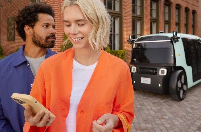 Two people look at a phone as a Zoox robotaxi waits nearby.